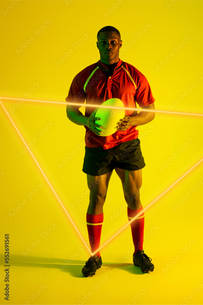 Confident african american male rugby player with ball by illuminated triangle on yellow background