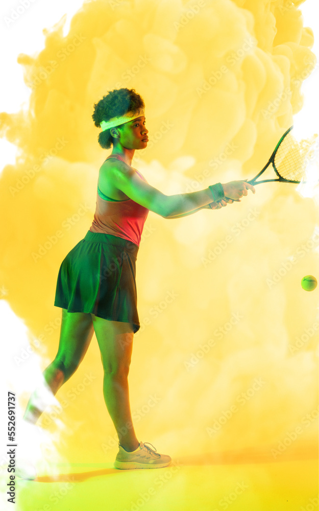 Side view of african american female player hitting ball with racket over yellow smoky background