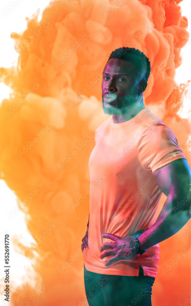Portrait of confident male athlete with hands on hip standing against orange smoky background