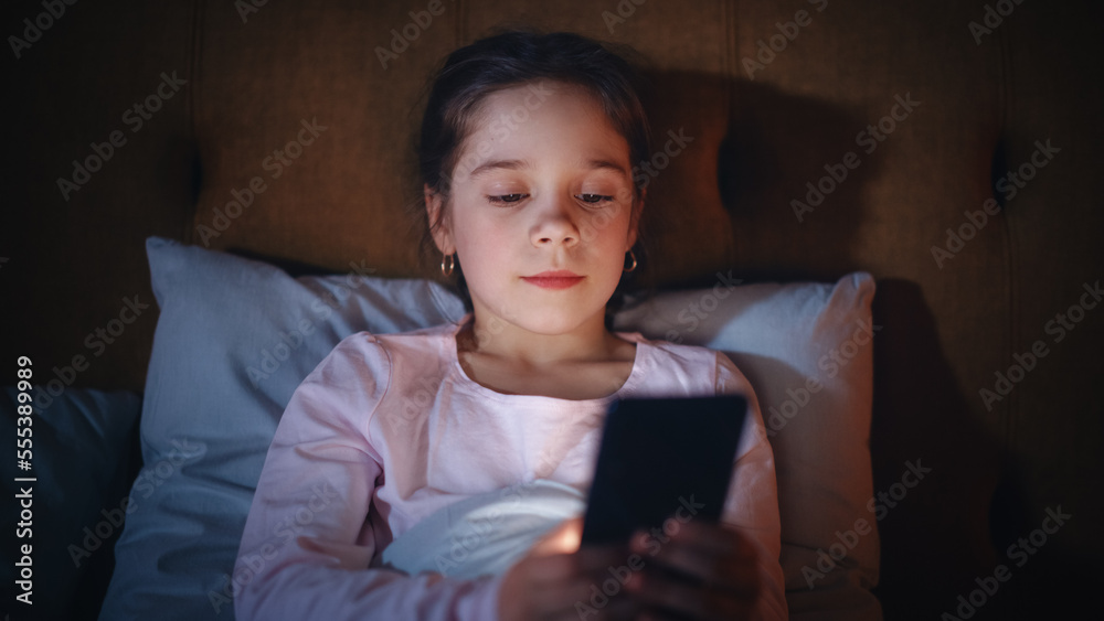 Small Happy Girl Watching Entertaining Videos on Smartphone in Bed Before Going to Sleep. Cute Kid L