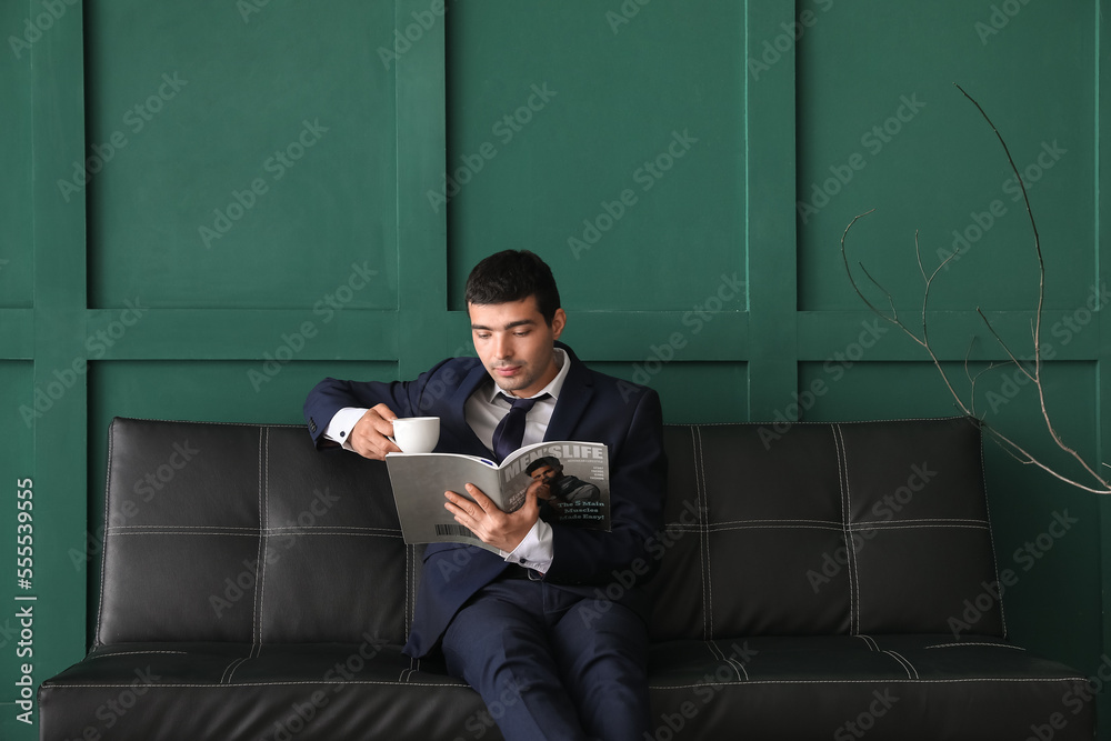年轻的商人拿着一杯咖啡在绿色墙壁附近的黑色沙发上阅读杂志