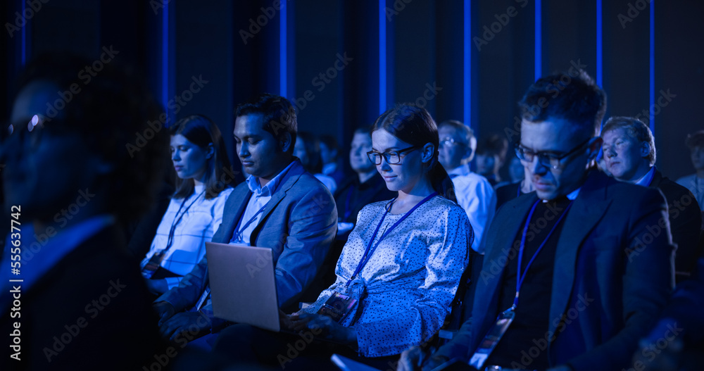 在一次技术会议上，女性坐在黑暗拥挤的礼堂里。年轻女性在使用笔记本电脑。