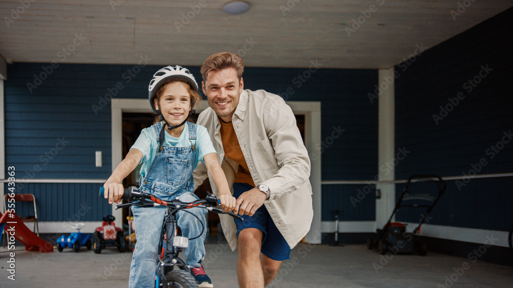 年轻的父亲在大房子的草坪上教女儿骑自行车。成功的女孩