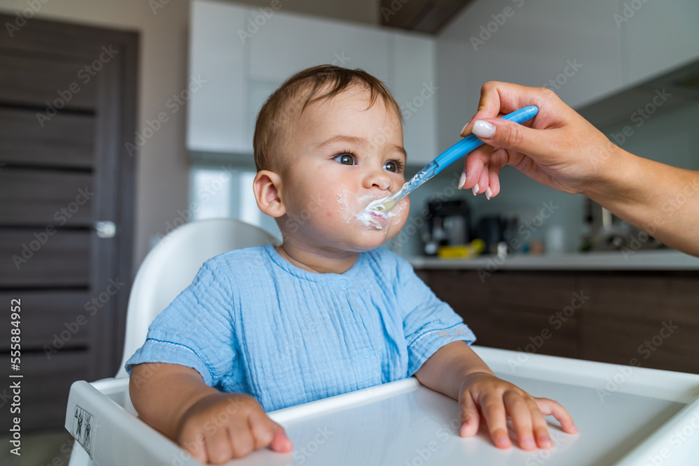 Mother feeding baby with spoon in kitchen. Baby eating. Portrait of happy young baby boy in high cha