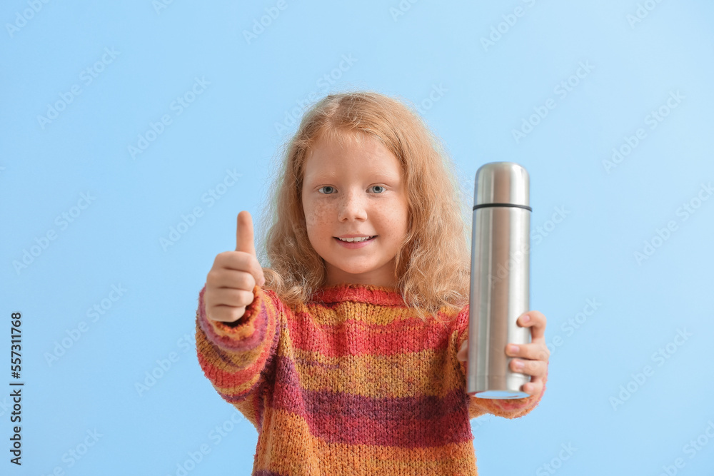 Little redhead girl with thermos showing thumb-up on blue background