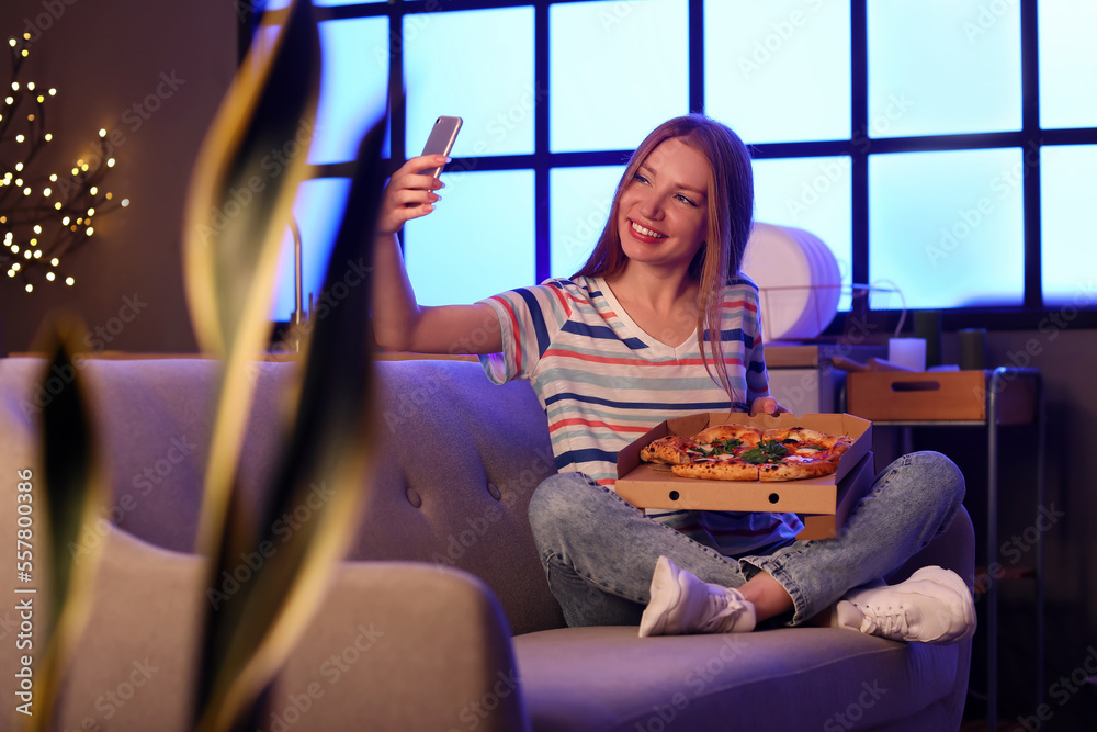 拿着美味披萨的年轻女子晚上在厨房沙发上自拍