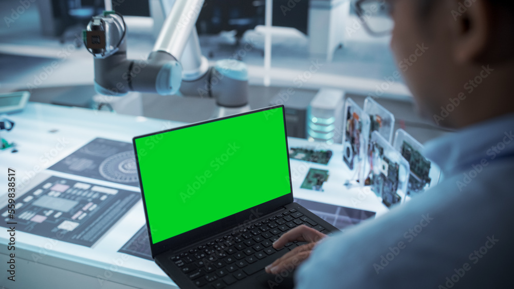 Over the Shoulder Shot of a Robotics Engineer Using a Laptop Computer with Green Screen Mock Up Disp