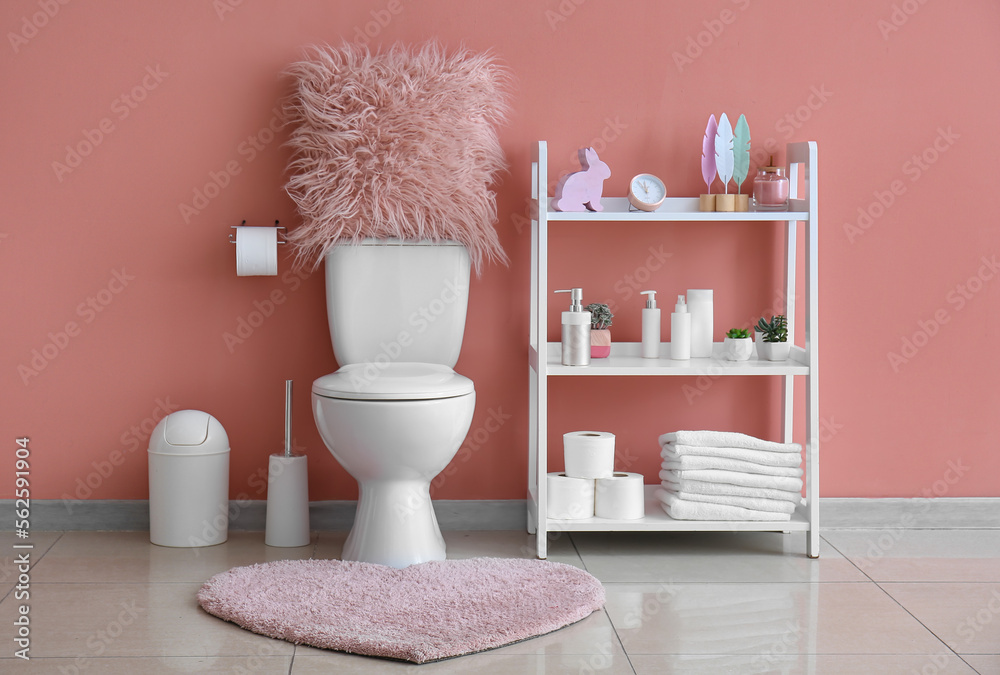 Interior of restroom with ceramic toilet bowl, rug and shelving unit near pink wall