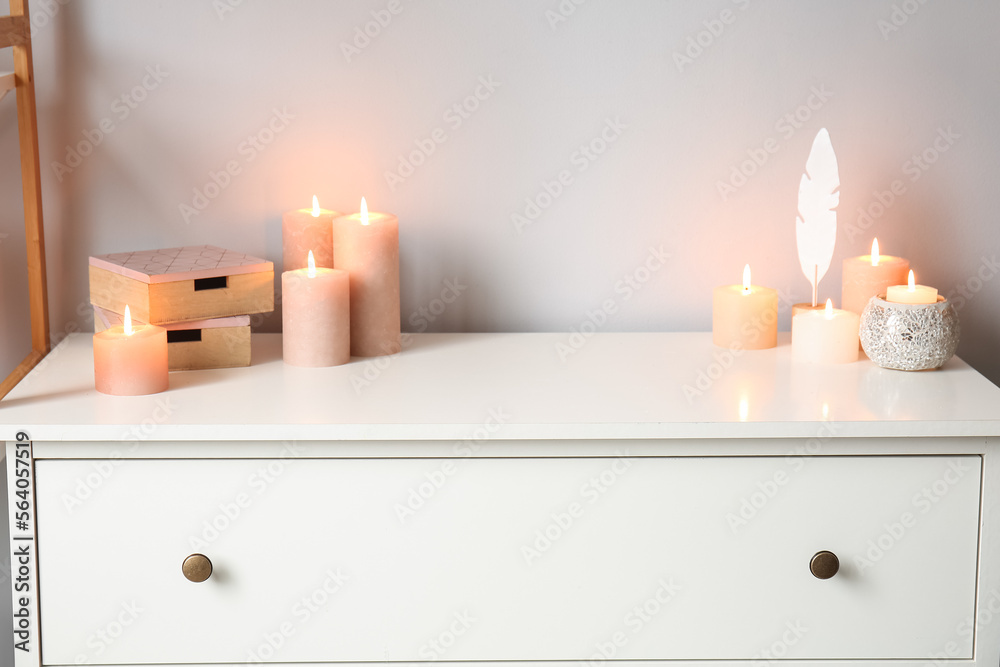 Burning candles, wooden boxes and decorative feather on drawer near white wall