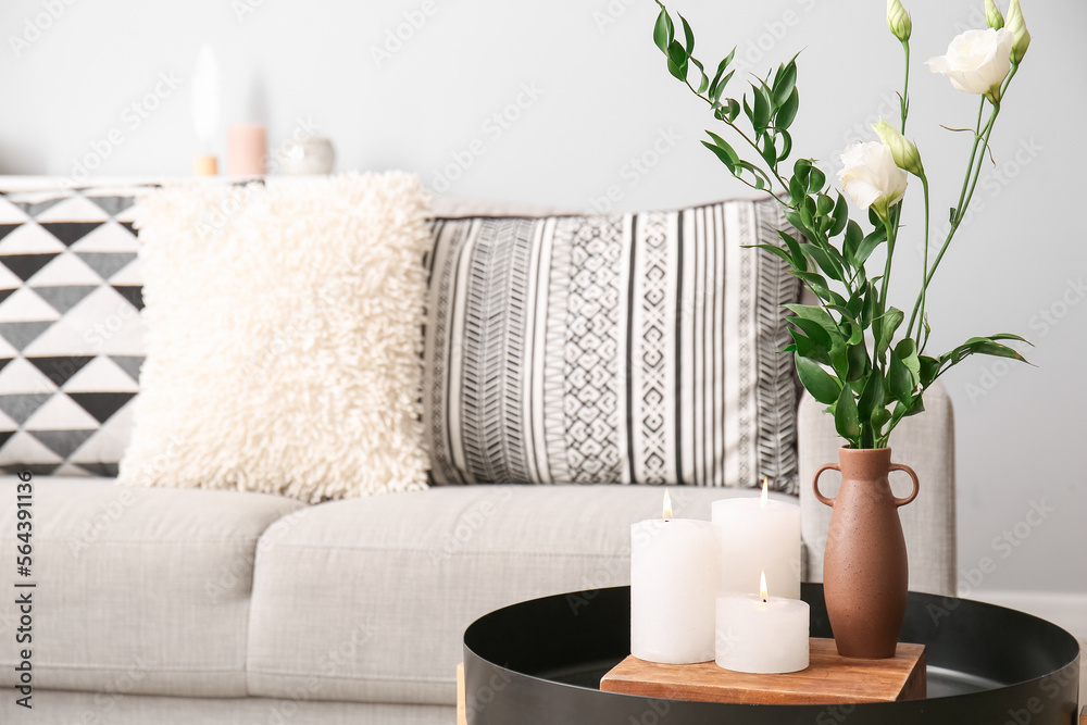 Burning candles and vase with eustoma and ruscus branches on end table near sofa in living room