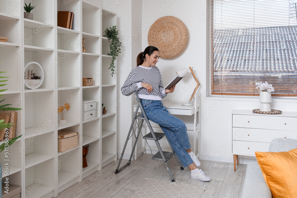 Young woman on stepladder reading book at home