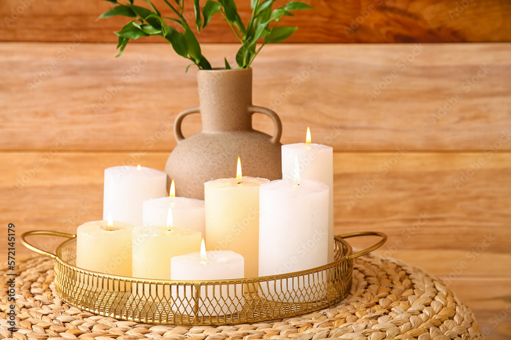 Tray with burning candles and plant branches in vase on pouf near wooden wall