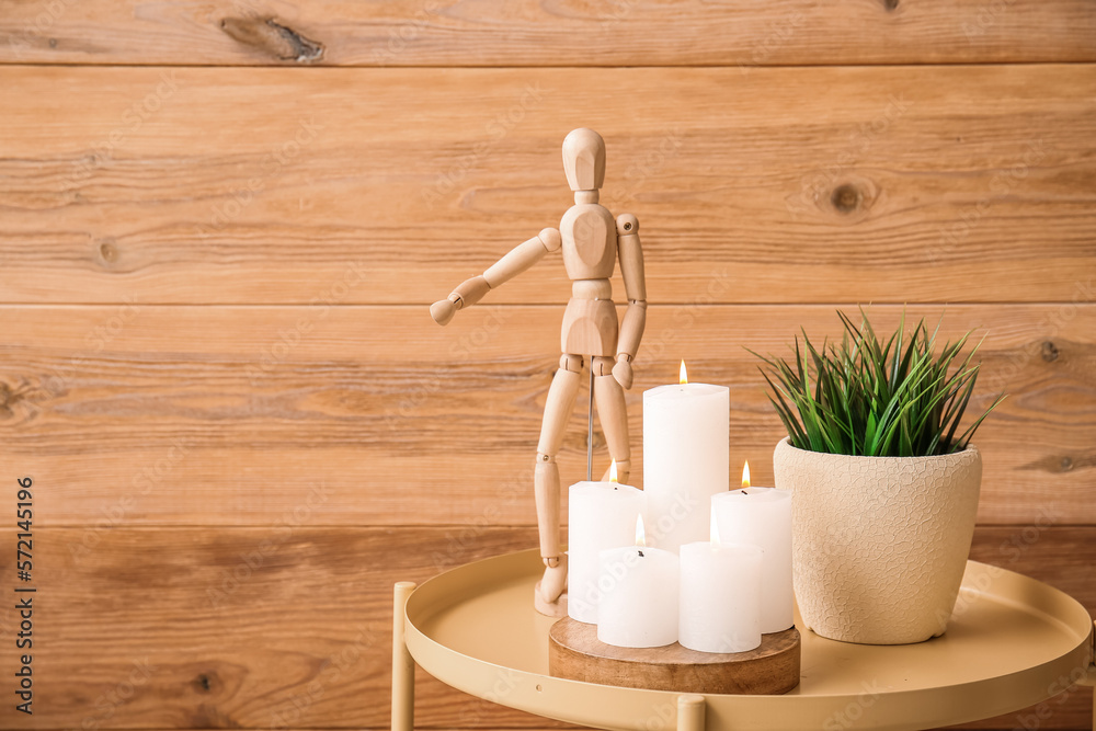 Burning candles, pot with grass and mannequin on table near wooden wall