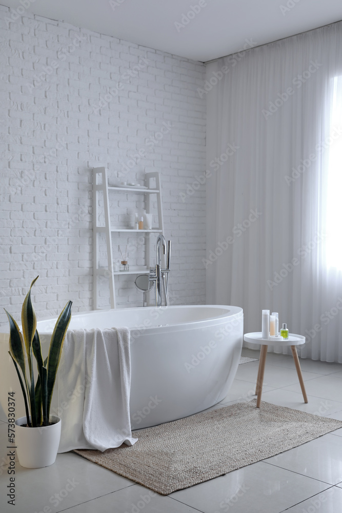 Interior of light bathroom with bathtub, table and shelving unit