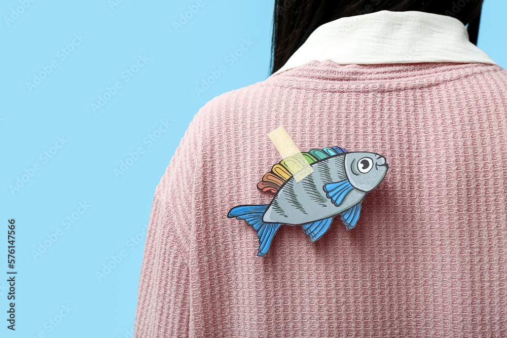 Paper fish on womans back against blue background, closeup. April Fools Day celebration