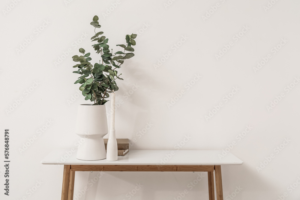 Vase with eucalyptus branches, burning candle and books on table near light wall