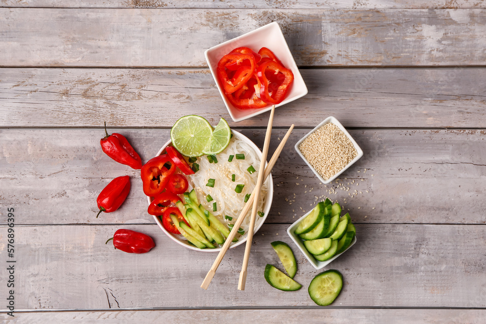 Composition with bowl of tasty rice noodles, vegetables and sesame seeds on light background