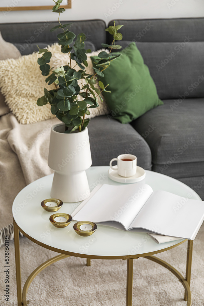 Vase with eucalyptus, cup of coffee, candles and magazine on table in living room