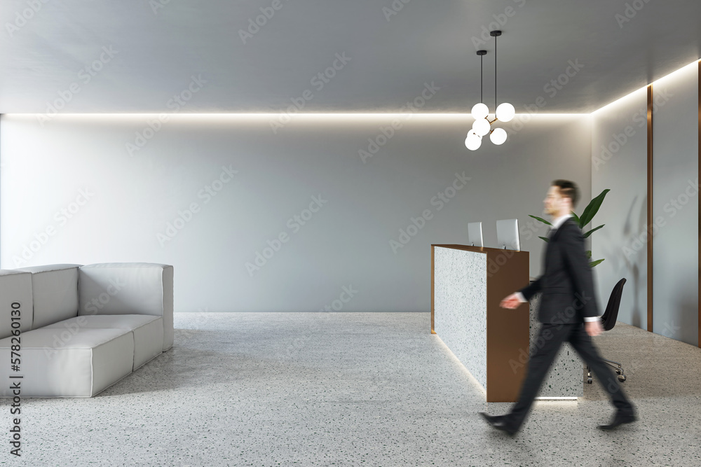 Businessman walking by modern reception desk in spacious office area with cozy grey sofa, concrete f