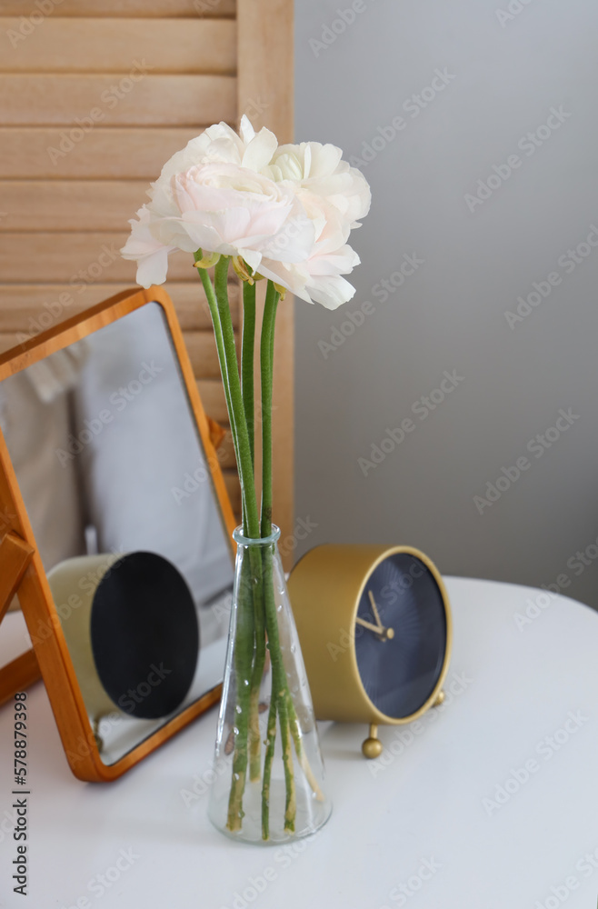 Vase with ranunculus flowers, alarm clock and mirror on table in bedroom