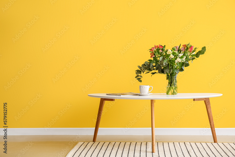 Vase with beautiful alstroemeria flowers, cup of coffee and magazine on table near yellow wall