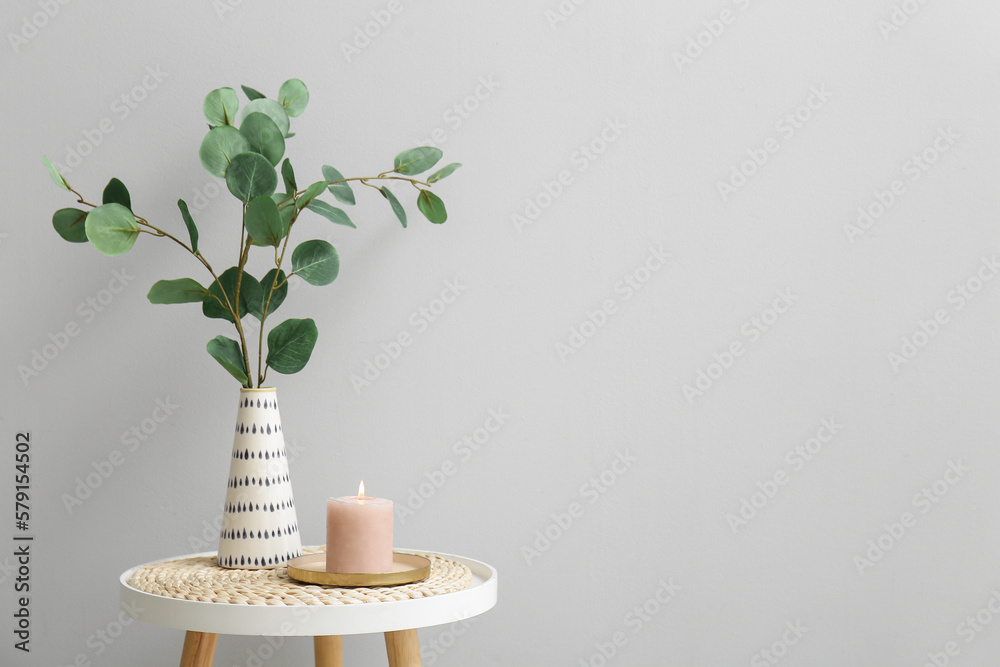 Burning candle and vase with eucalyptus branches on table near grey wall