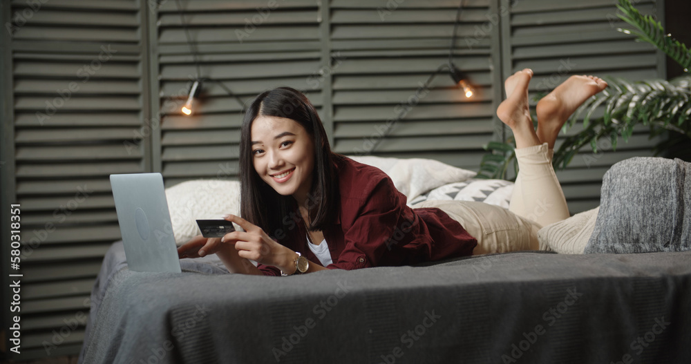 Young Asian student spending time on online shopping, lying on large bed with her laptop, keying in 