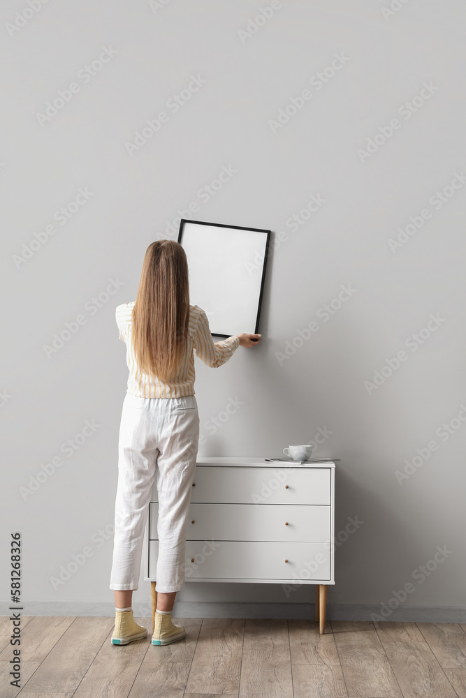 Young woman hanging blank frame on grey wall at home