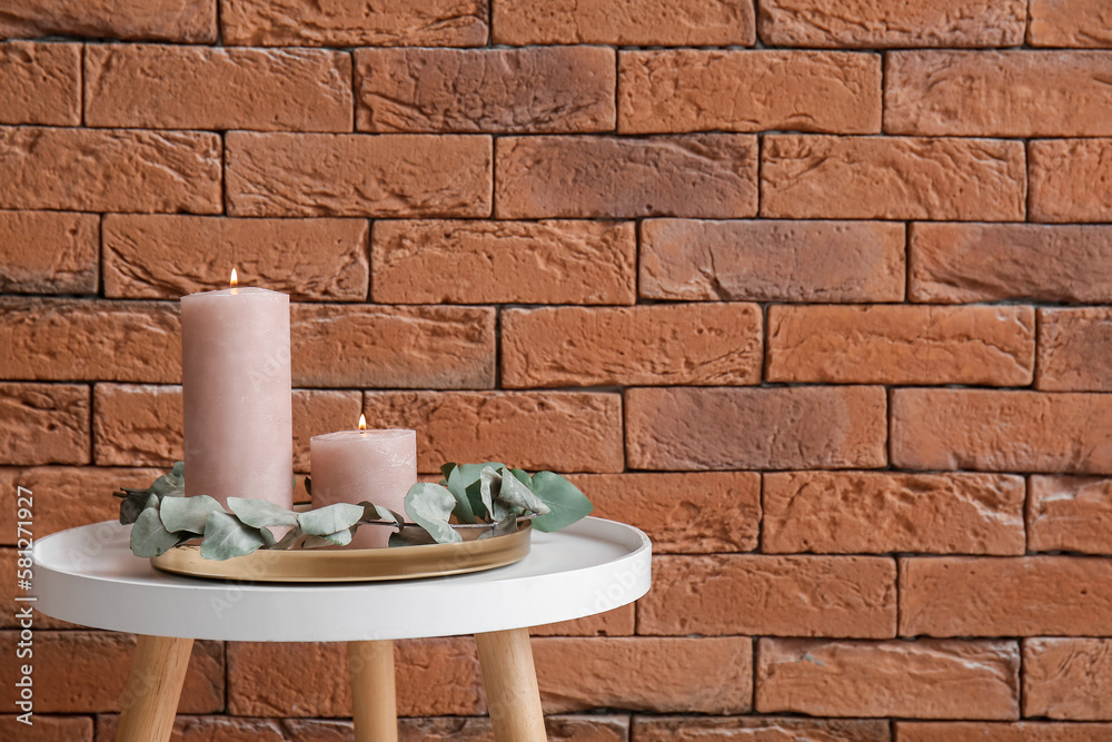 Burning candles and eucalyptus branch on table near brown brick wall