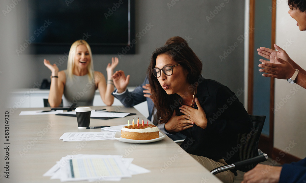 Business woman celebrating her birthday with her colleagues, blowing out candles on a cake in the of