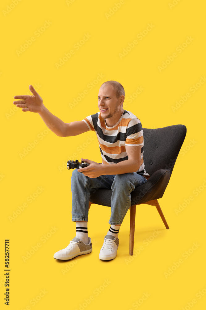 Handsome man playing video game in black armchair on yellow background