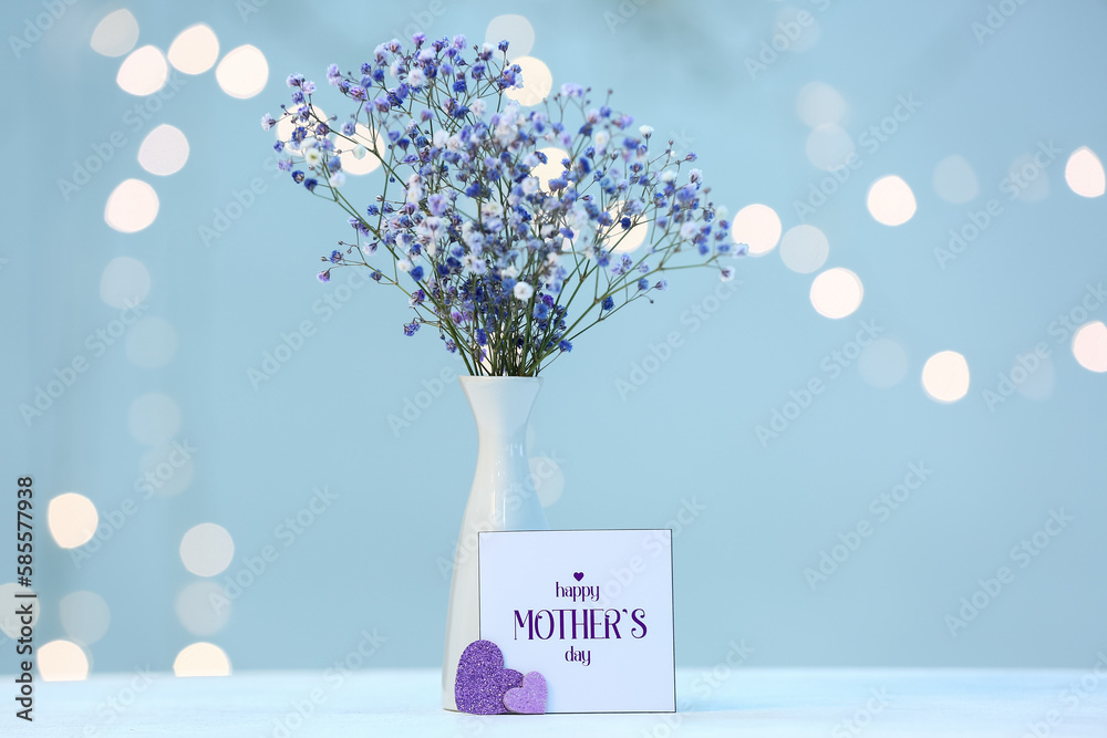 Card with text HAPPY MOTHERS DAY and vase of beautiful gypsophila flowers on table against blurred 