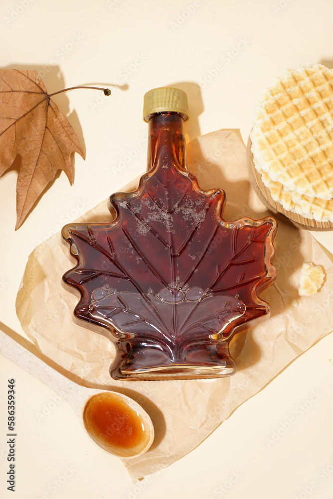 Bottle and spoon of tasty maple syrup on beige background
