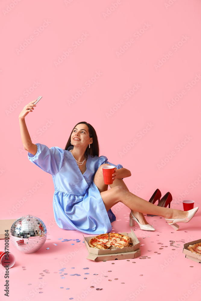 Young woman with cup, tasty pizza and disco ball taking selfie on pink background