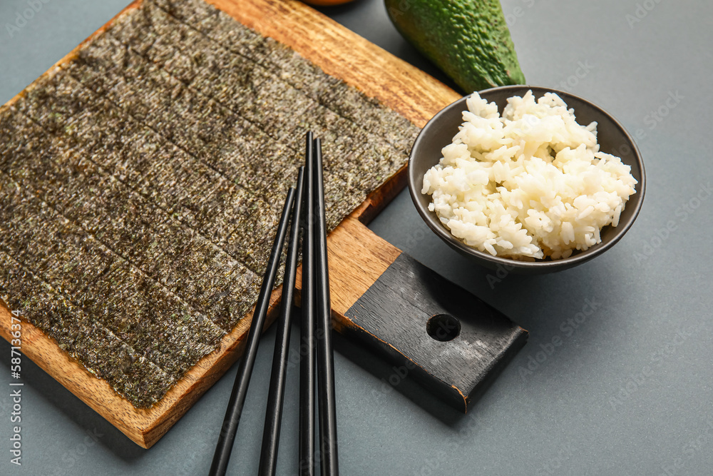 Wooden board with nori sheet, bowl of rice and chopsticks on dark background, closeup