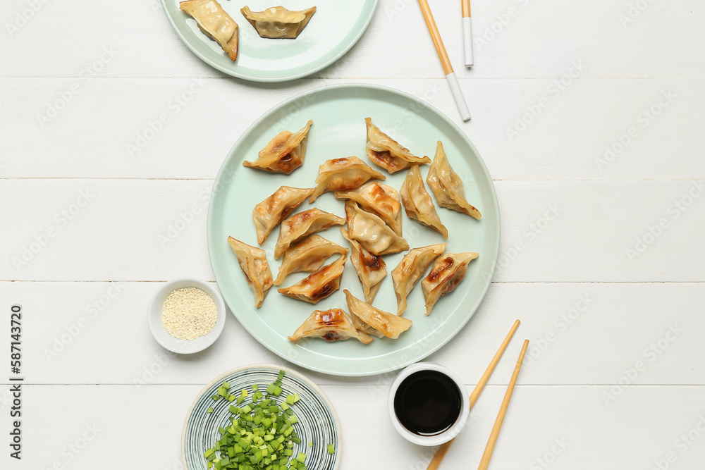 Plates with tasty Chinese jiaozi, onion and sauce on white wooden background