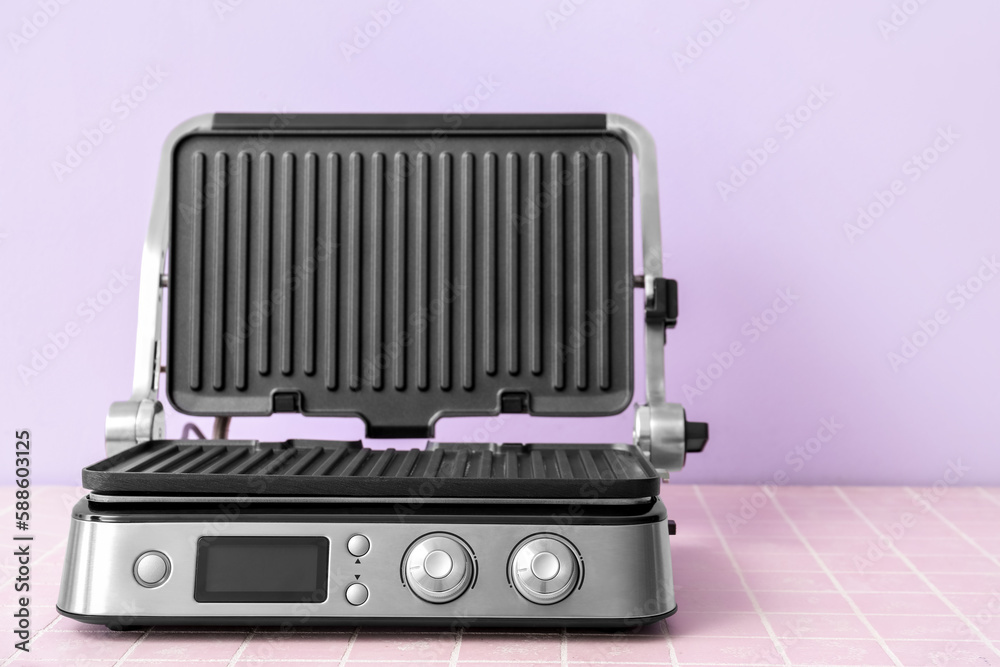 Modern electric grill with open lid on pink tile table against lilac background