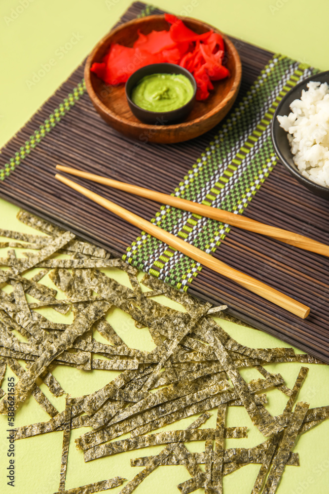 Bamboo mat with ginger, wasabi and cut nori sheets on green background, closeup