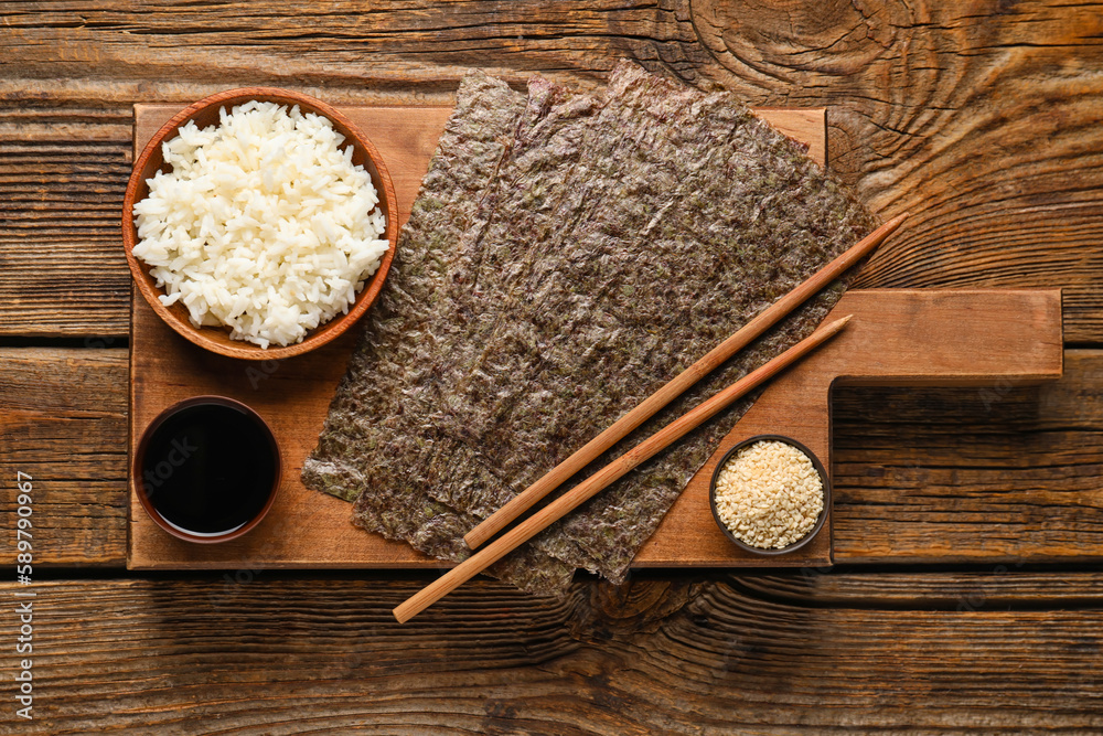 Board with nori sheets, rice, sesame seeds and sauce on wooden background