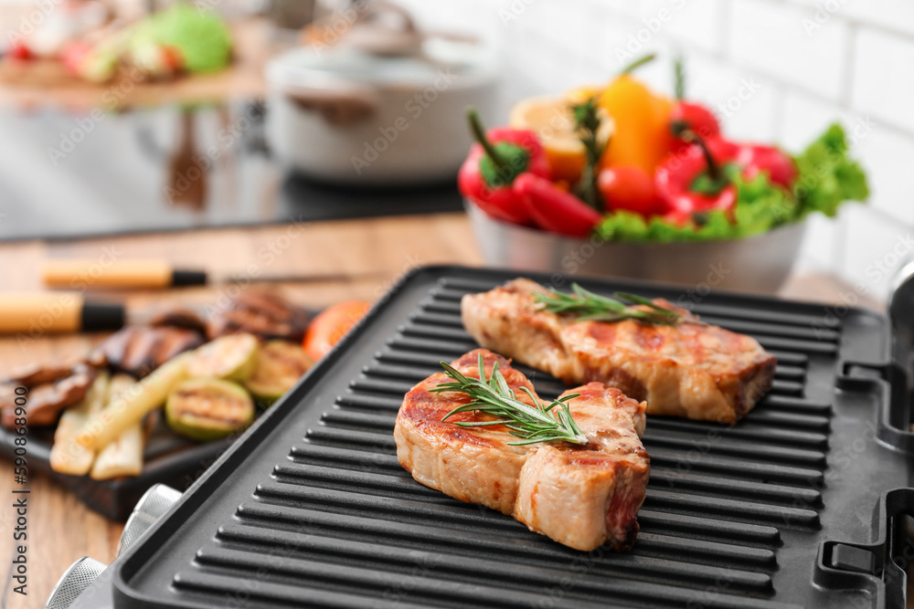 Modern electric grill with tasty steaks on table near light brick wall, closeup
