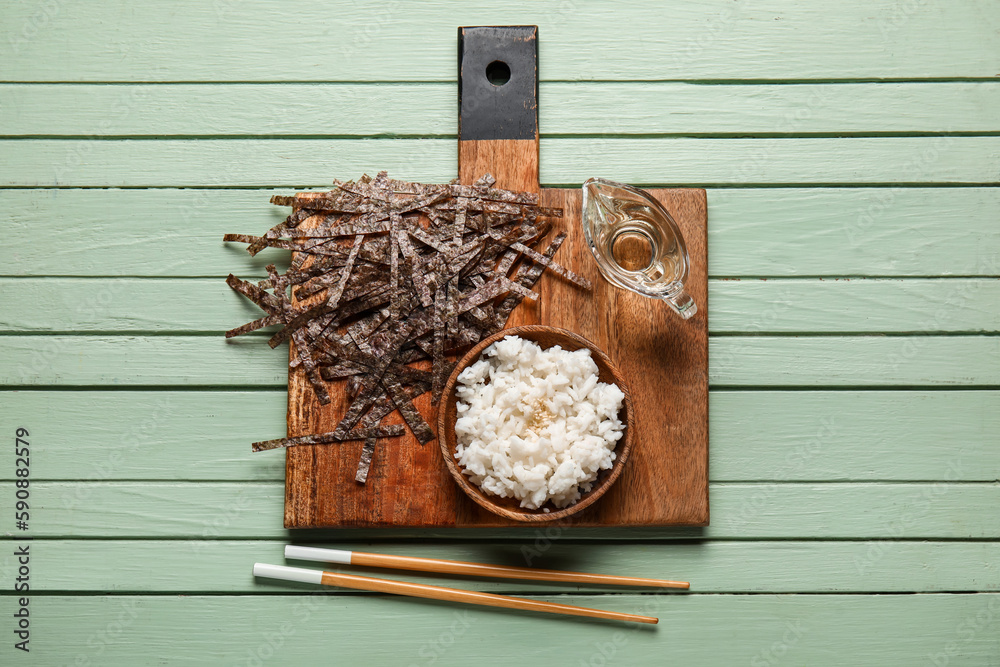 Cutting board with plate of boiled rice, vinegar, cut nori sheets and chopsticks on color wooden bac