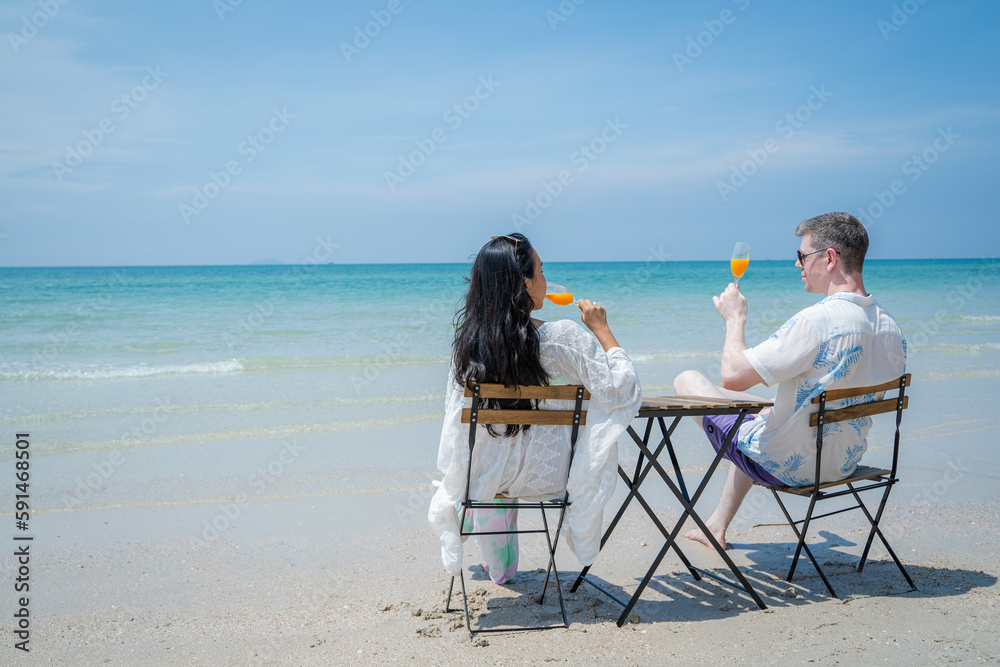 Smiling couple enjoying drinks orange juice while resting on comfortable chairs by sea beach,Summer 