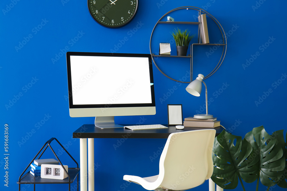 Workplace with computer, books and lamp near blue wall in office