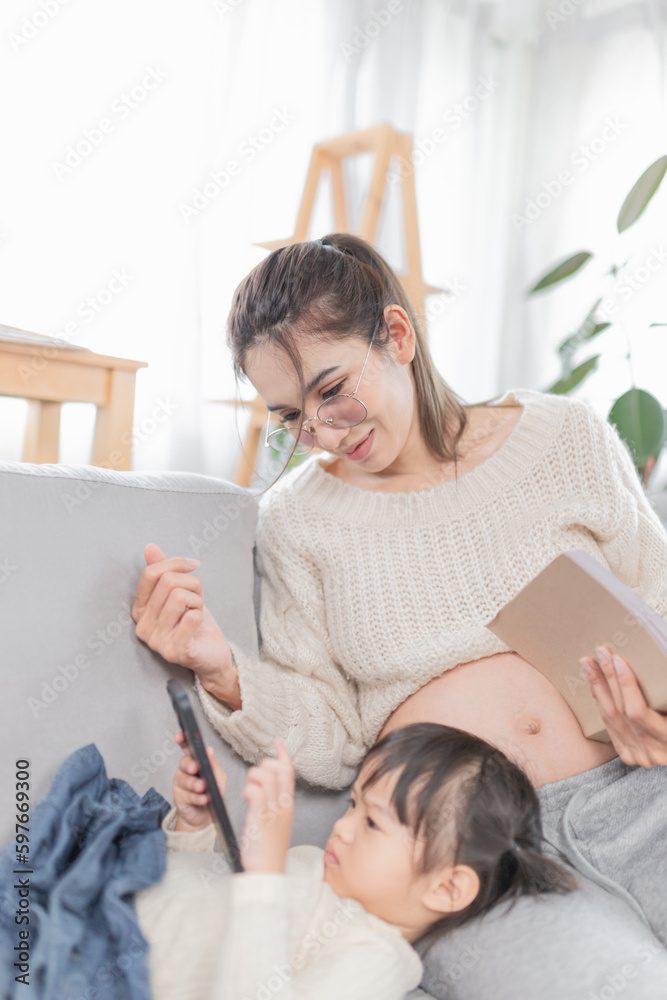 close up mother, asian female and her daughter relax on sofa, pregnant woman reading a book, a girl 