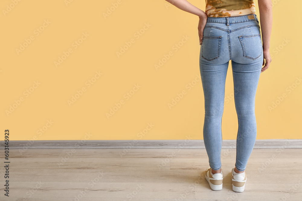 Young woman in stylish jeans near yellow wall, back view