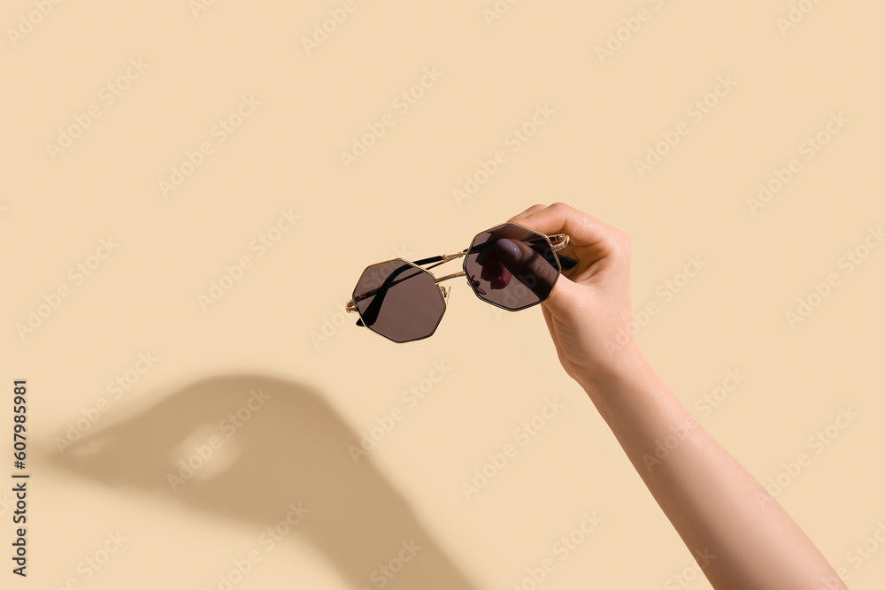 Female hand holding stylish sunglasses on beige background