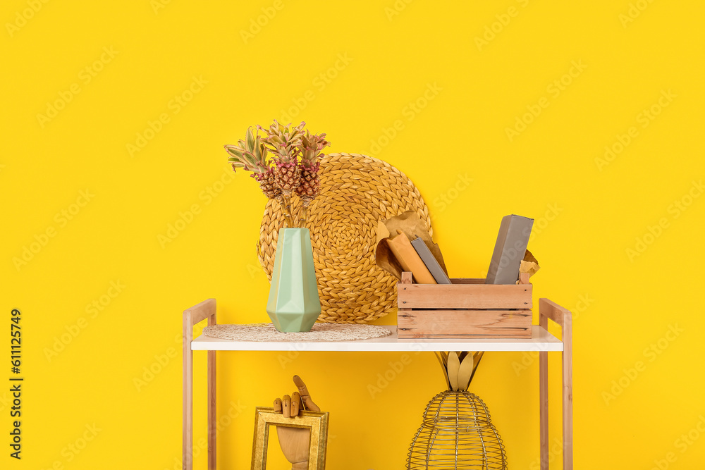 Vase with baby pineapples, books and decor on shelves near yellow wall