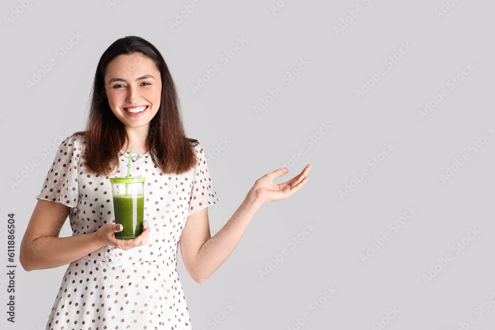 Young woman with glass of vegetable juice showing something on grey background