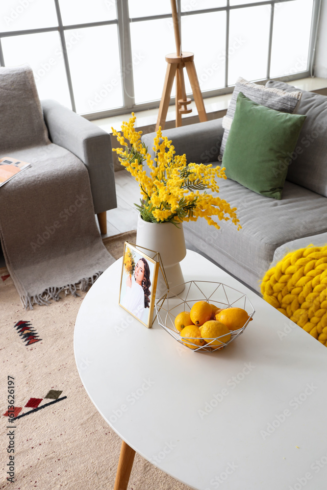 Vase with mimosa flowers, lemons in basket and womans photo on coffee table in living room