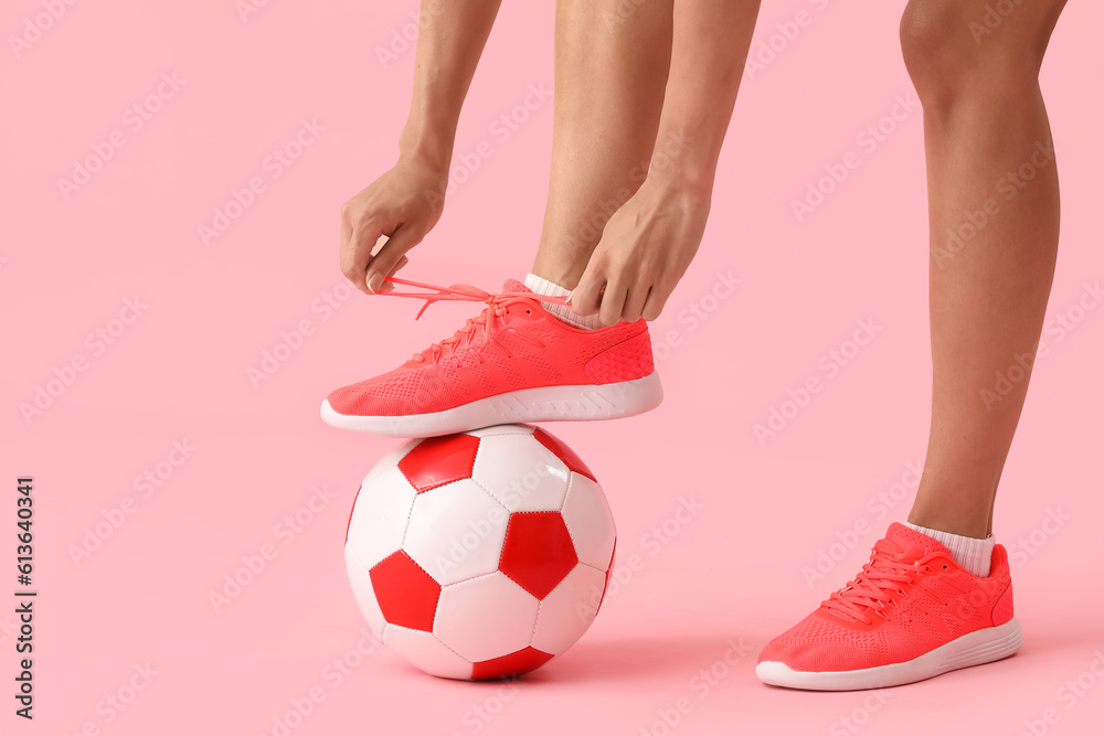 Legs of young woman with soccer ball tying shoelaces on pink background